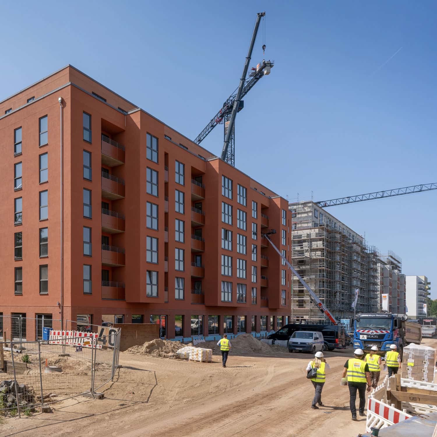 Auf der Baustelle Neues Gartenfeld stehen mehrere mehrgeschossige Wohnneubauten, teils mit rotbrauner Fassade, teils noch im Gerüst. Im Vordergrund sind eine Baustellenstraße, Baufahrzeuge und mehrere Personen mit Schutzhelmen und Warnwesten zu sehen.